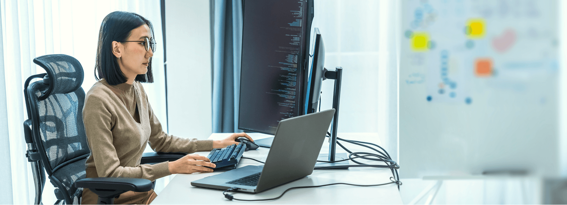 A woman sitting at a desk in an office, working on a desktop computer with code displayed on the monitor, while using a keyboard and mouse, with a laptop open beside her.